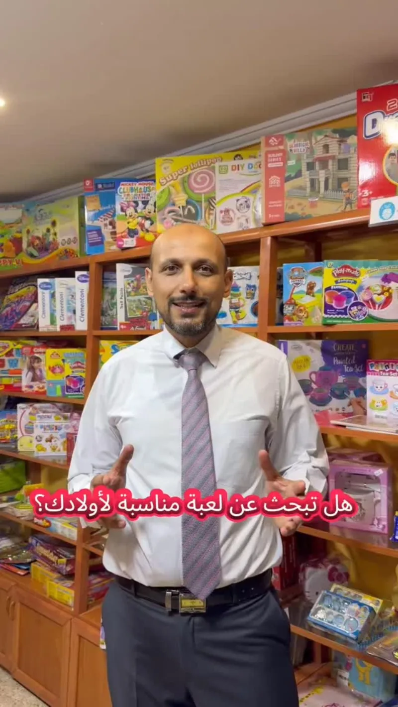 رجل بقميص وربطة عنق يقف أمام رفوف مليئة بعلب الألعاب.